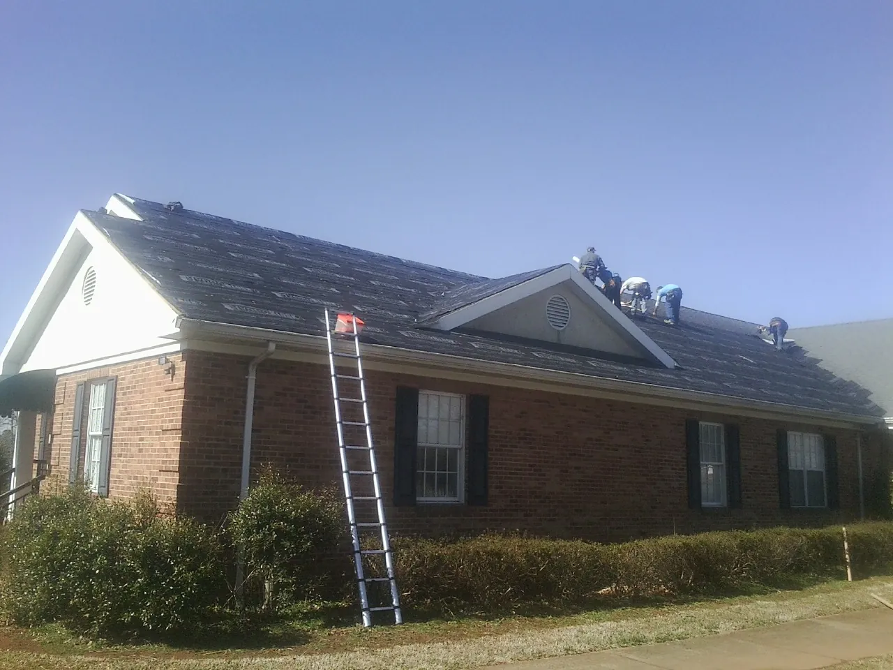 Crew tearing shingles off a brick ranch roof with a ladder set — Emergency Roof Repair work by our crew in Spruce Creek