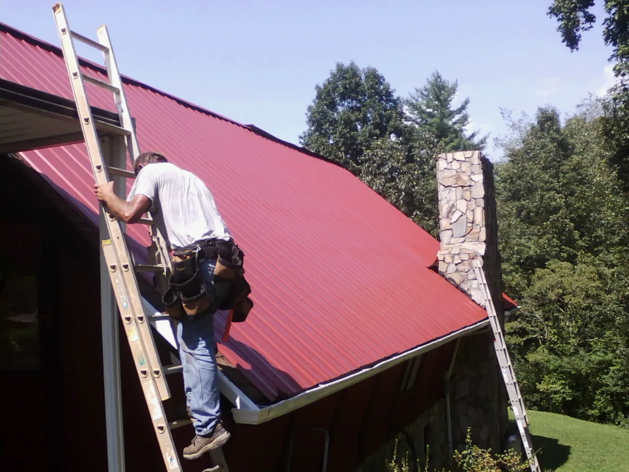 Roofer on a ladder beside a red metal roof and stone chimney — Soffit & Fascia Repair by Roofing Brothers in Spruce Creek